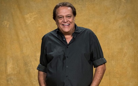 Dennis Carvalho sorri com as mãos no bolso usando camisa preta em frente a uma parede amarela-mostarda