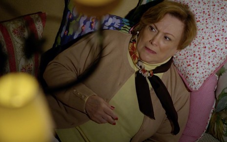 A atriz Nicette Bruno como Iná tem uma expressão de preocupação e está deitada em um sofá cheia de almofadas em cena de A Vida da Gente