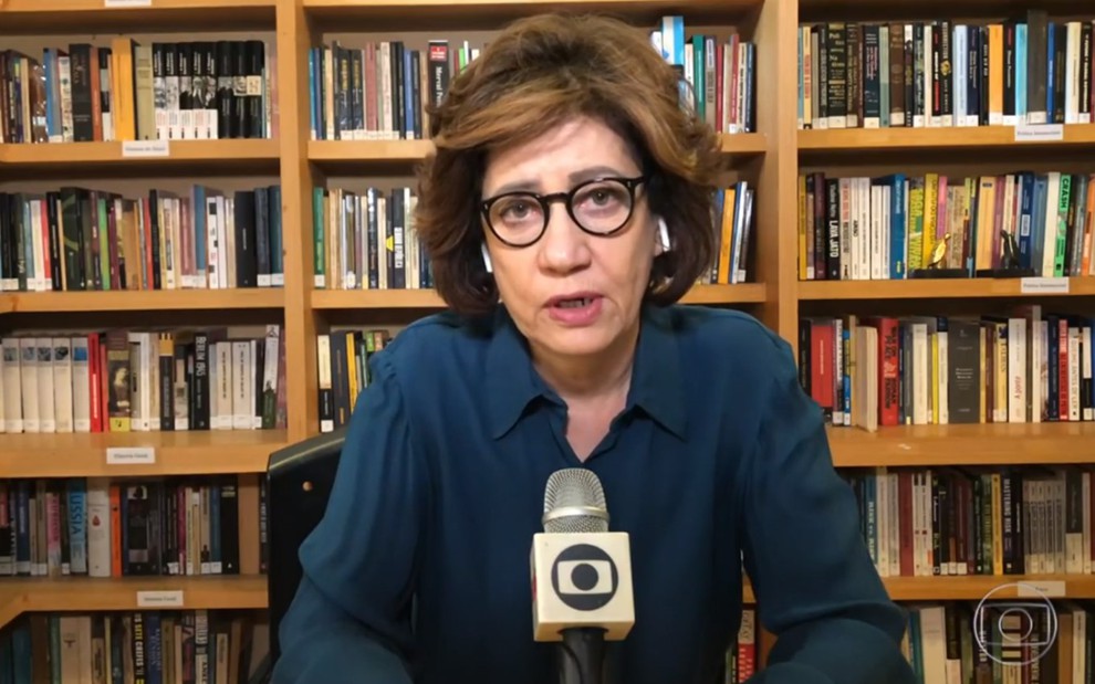 Miriam Leitão de camisa azul e óculos, com uma estante cheia de livros ao fundo