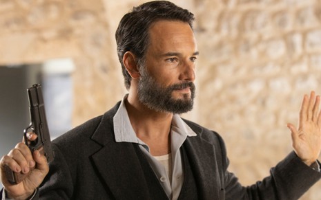 Rodrigo Santoro levanta as mãos para o alto em sinal de redenção enquanto segura uma pistola na mão direita