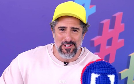 Mion olha para frente, veste blusa rosa, segura um bastão com o logo da Globo e usa boné amarelo