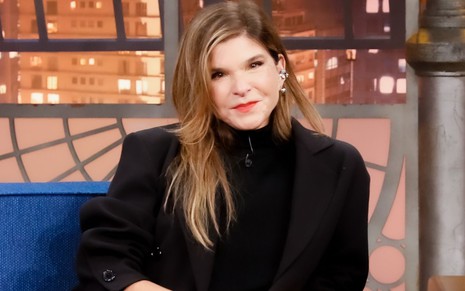 Cristiana Oliveira, vestida de preto, sorri para foto em cenário do programa The Noite, do SBT