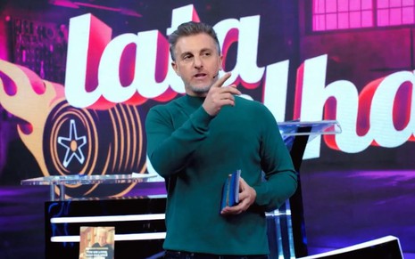 Luciano Huck aponta com o indicador, como se fizesse o número 1, com a marca do Lata Velha no telão ao fundo