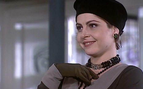 Drica Moraes, caracterizada como Marcela, exibe um sorriso de vitória; uma das mãos estão apontadas para o peito. Ela veste um chapéu, vestido preto --com gola cinza--, luva marrom e colar de pedras pretas.