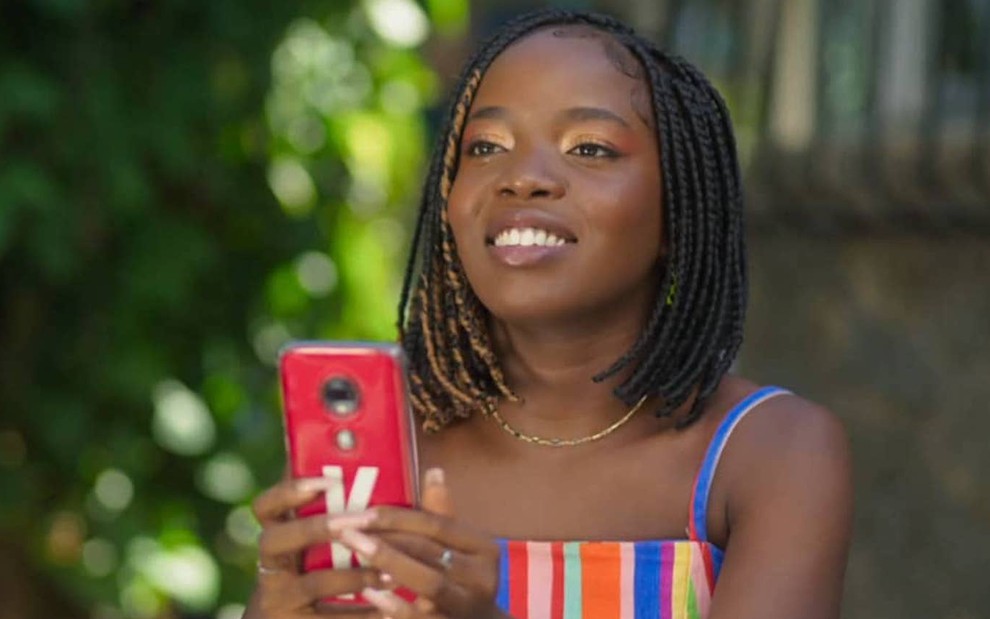 Clara Moneke segura celular e sorri em cena de Vai na Fé