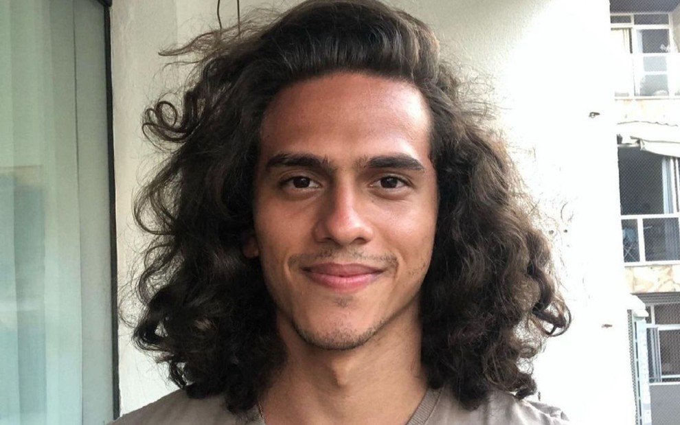 Ruan Aguiar está de cabelo solto, na altura do ombro; ele está em sacada de apartamento