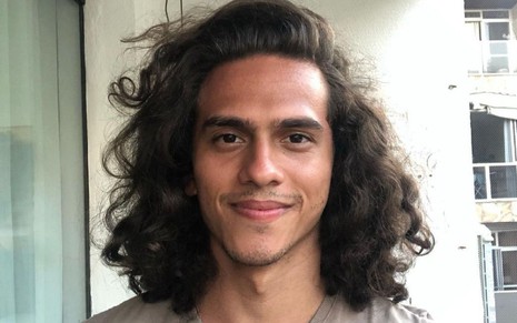 Ruan Aguiar está de cabelo solto, na altura do ombro; ele está em sacada de apartamento