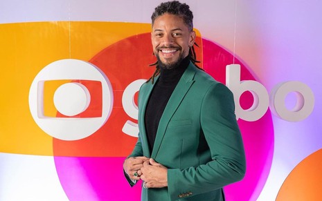 Paulo Lessa está usando um blazer verde militar com uma blusa de gola preta; ele posa em frente ao logo da Globo com o nome da emissora