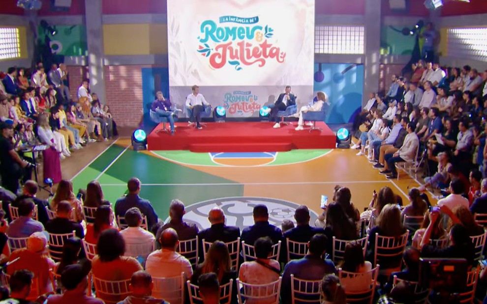 Plateia cerca um pequeno palco com quatro pessoas sentadas abaixo do logotipo da novela A Infância de Romeu e Julieta