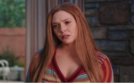 Wanda (Elizabeth Olsen) com blusa vermelha colorida com cabelo solto nos ombros com cenário de uma casa ao fundo em cena de WandaVision