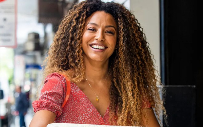 A atriz Sheron Menezzes como Sol em Vai na Fé; ela está olhando para frente e sorrindo para a foto enquanto segura uma caixa de isopor