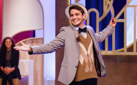 Vestido com paletó, colete, gravata-borboleta e chapéu, Victor Lamoglia abre os braços e força um sorriso no cenário do M.E.M.E. da Comédia