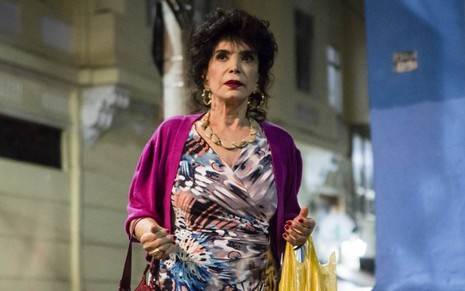 A atriz Lady Francisco (1935-2019) em cena de Totalmente Demais (2015), com expressão séria, vestido estampado, casaco roxo