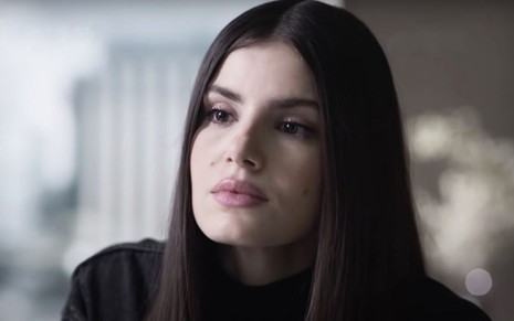 Camila Queiroz como Angel. Ela usa um vestido preto, um cabelo longo e olha de forma sedutora para a câmera