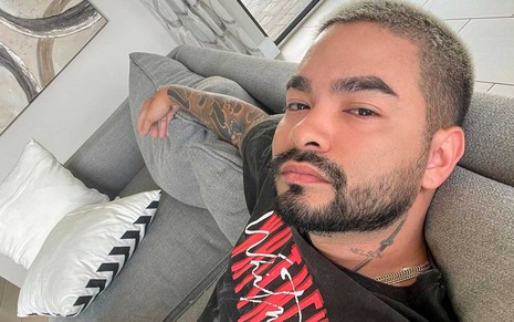 O apresentador Yudi Tamashiro, com uma camisa preta com letras vermelhas, jogado em um sofá cinza