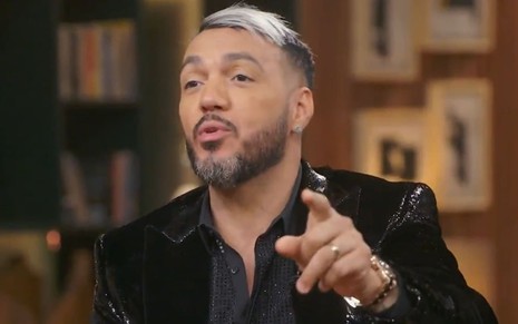 Belo levanta a mão esquerda, e é possível ver a aliança de ouro em seu dedo anelar. Ele usa um terno preto e está falando