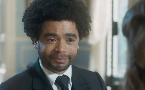 Diogo Almeida caracterizado como Orlando; ele usa um terno preto e sorri, com os olhos cheios de lágrimas, em cena de Amor Perfeito