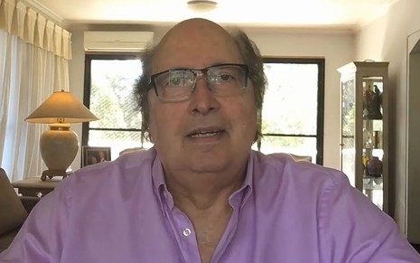 Luciano Callegari usa uma camisa rosa e está de óculos de grau; ao fundo há uma grande janela
