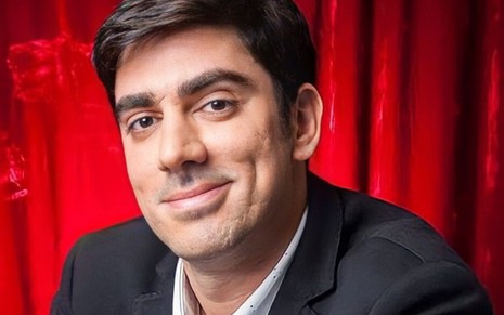 Marcelo Adnet está com um sorriso discreto, em frente a um fundo de uma cortina vermelha