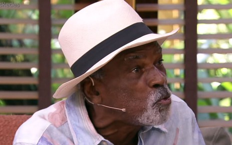 Antônio Pitanga sentado no sofá do É de Casa, usando chapéu branco com faixa preta