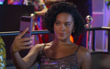 Bella Campos está com o celular erguido em uma das mãos, como Maria de Fátima Acioli em Vale Tudo