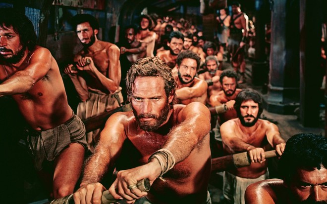 Sem camisa, Charlton Heston rema em um navio juntamente com outros personagens