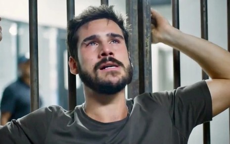 Nicolas Prattes tem expressão de desespero enquanto agarra barras de prisão em cena de Mania de Você