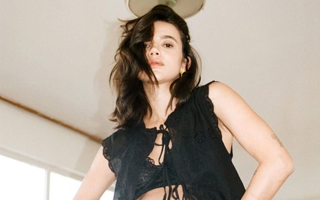 Manu Gavassi está com cabelos soltos e usa blusa preta aberta com top; ela coloca a mão na barriga de grávida