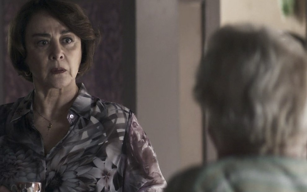 A atriz Nívea Maria segura uma xícara caracterizada como a Evelina em cena de A Dona do Pedaço, com Suely Franco, a Marlene, de costas, olhando para ela